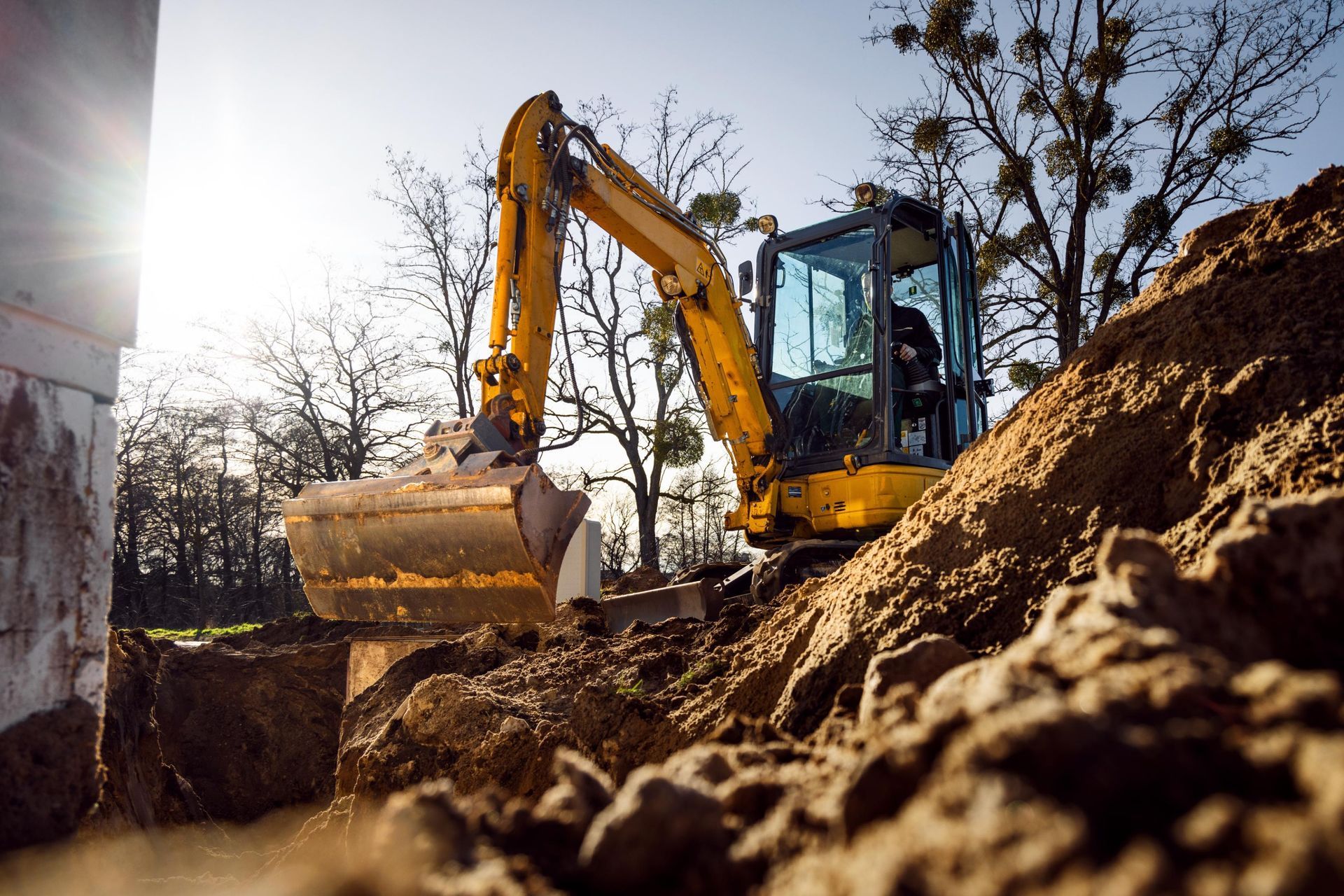 Yellow excavator digging dirt on a construction site beside a mound of earth and bare trees