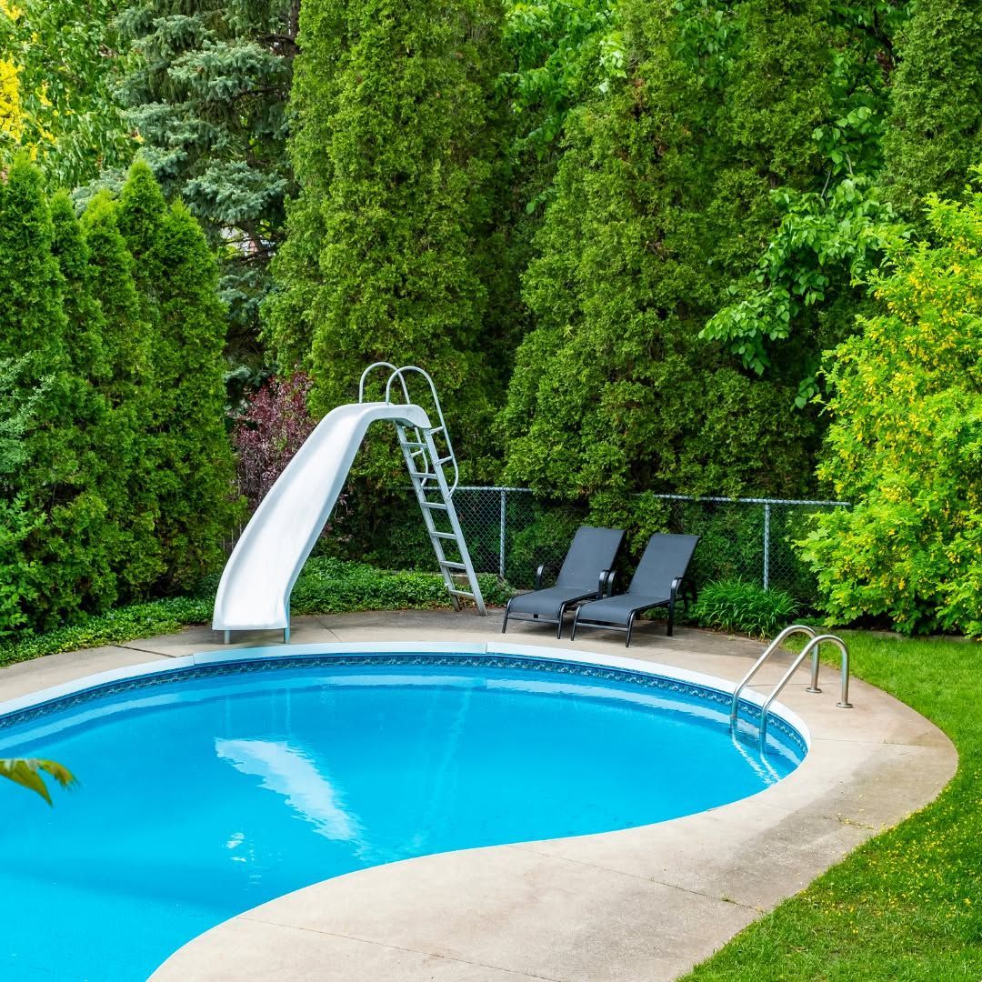 A backyard swimming pool with a slide, two lounge chairs, and a metal fence, surrounded by tall evergreen trees.