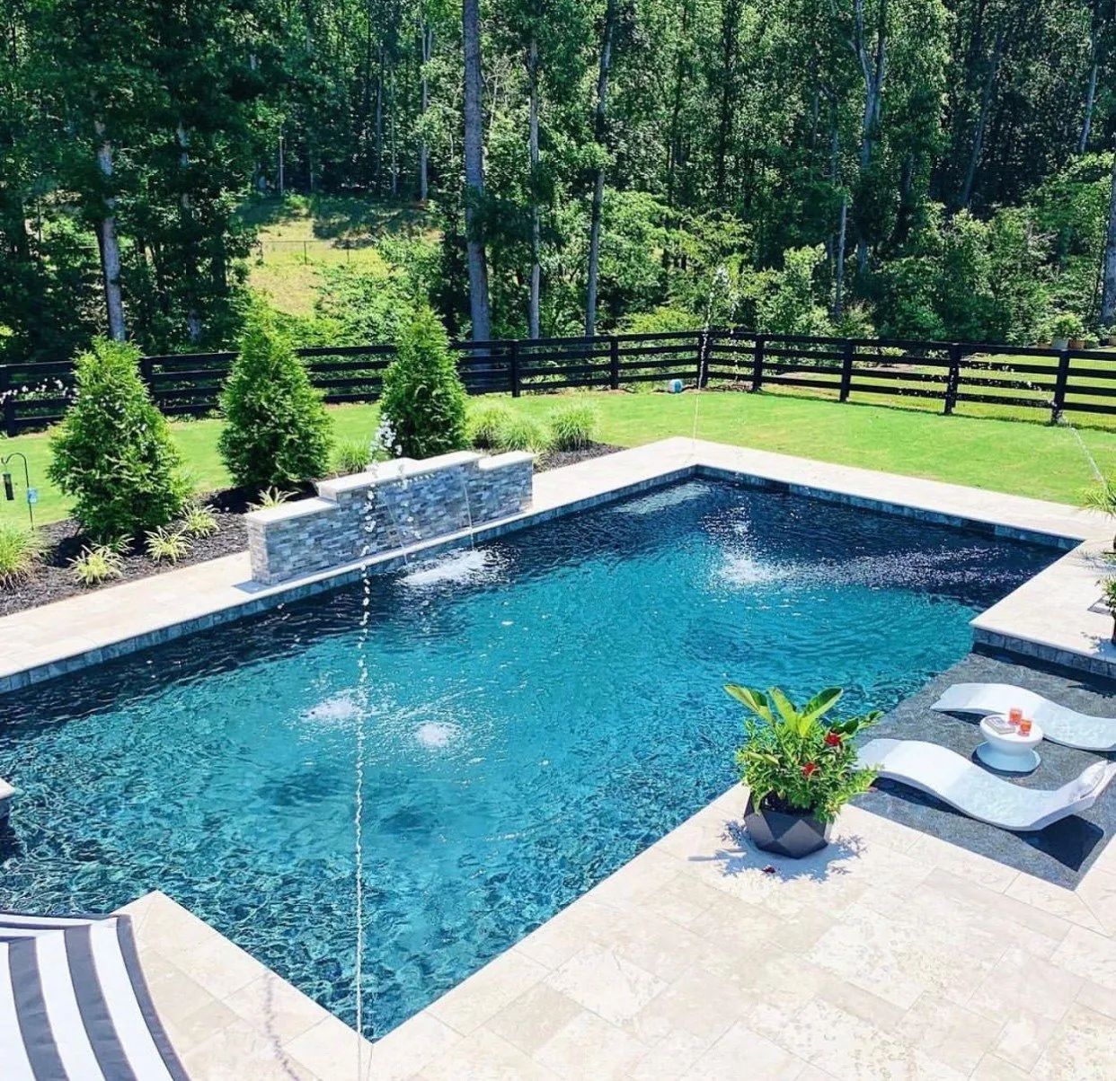 Backyard pool with fountain, stone patio, lounge chair, and green lawn with trees behind a wooden fence