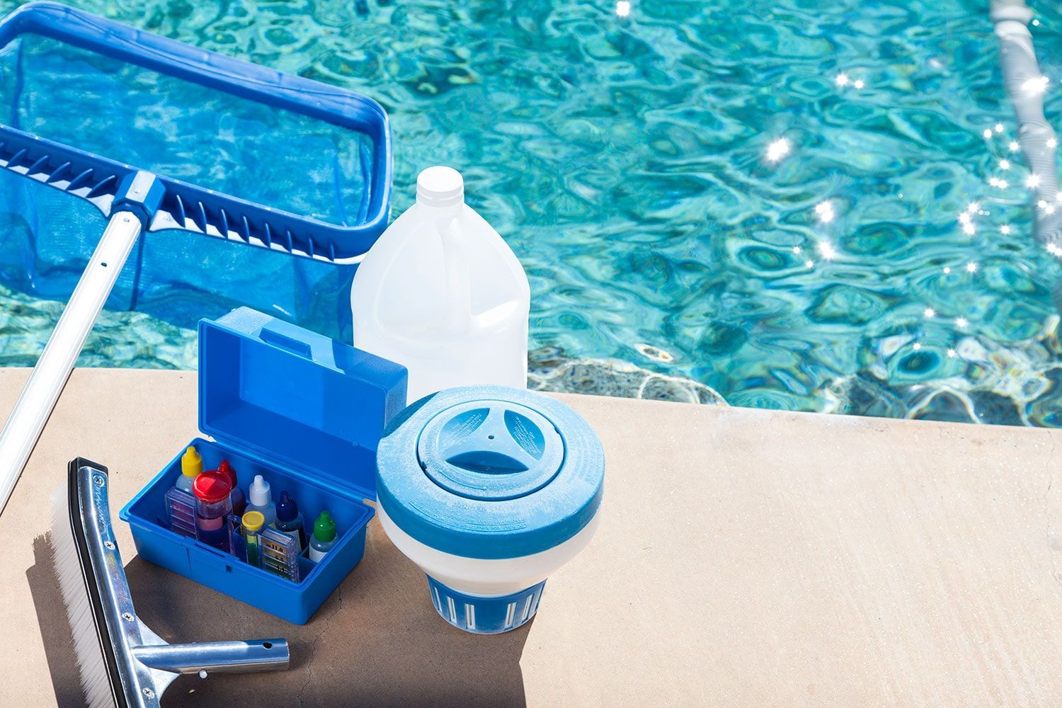 Pool cleaning supplies, including a skimmer net, test kit, chemicals, and a floating chlorine dispenser, by a pool.