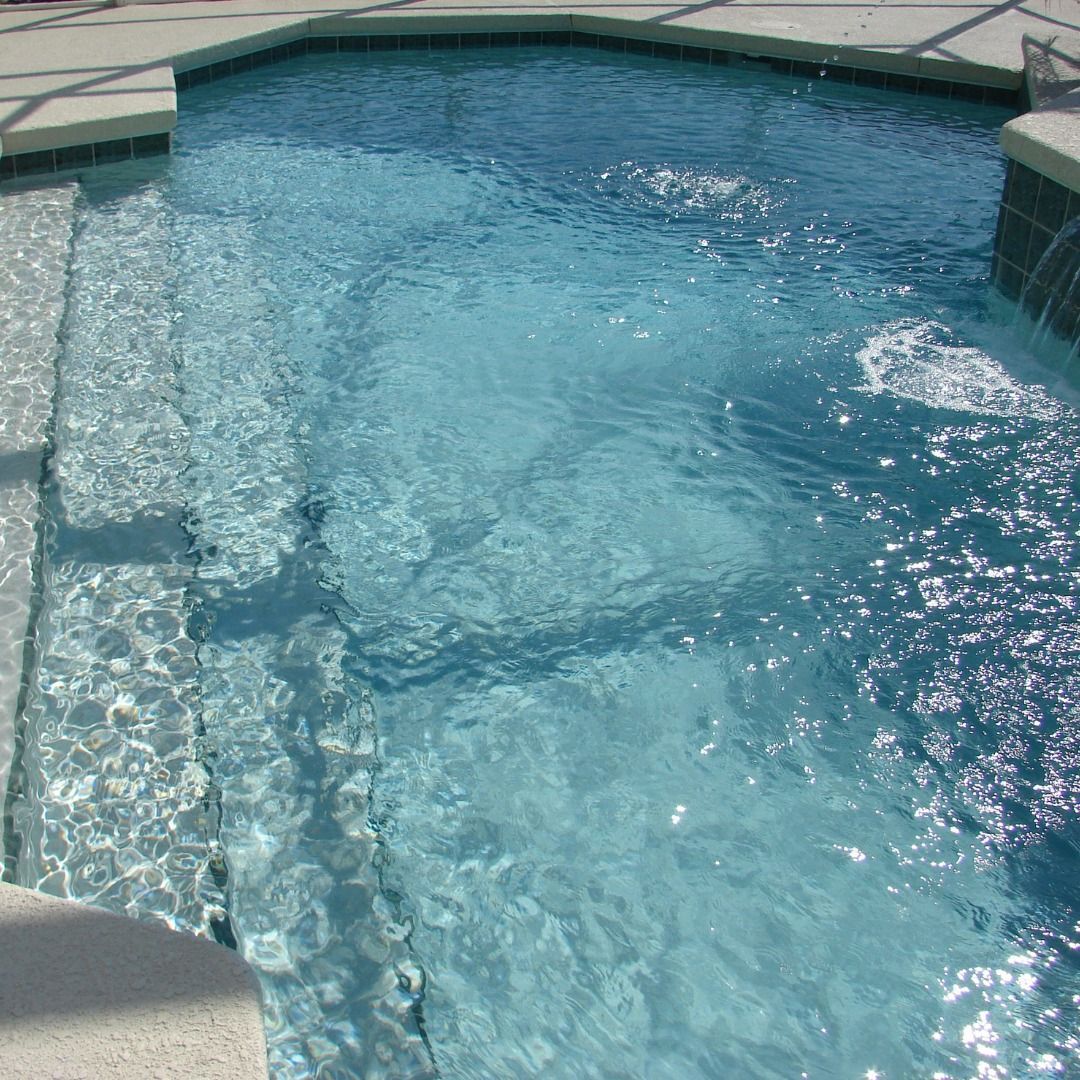 A rectangular swimming pool with blue tiles, surrounded by a stone border and lush green plants.