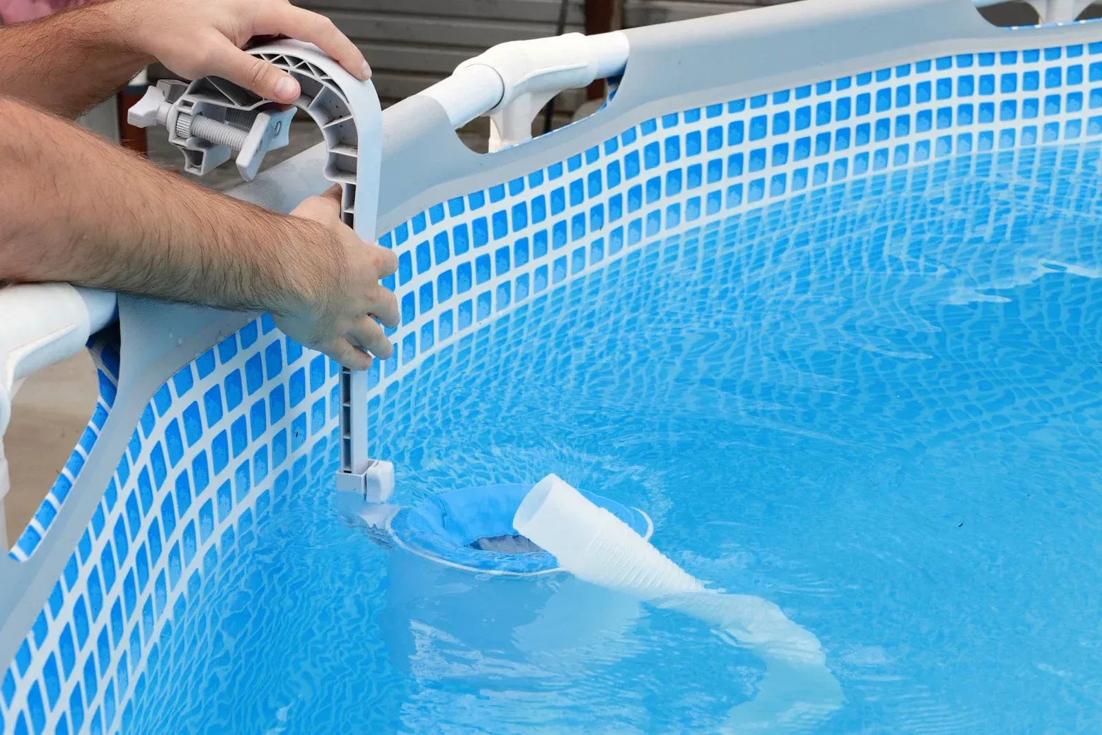 Hands installing a white swimming pool skimmer attachment onto the frame of an above-ground pool.