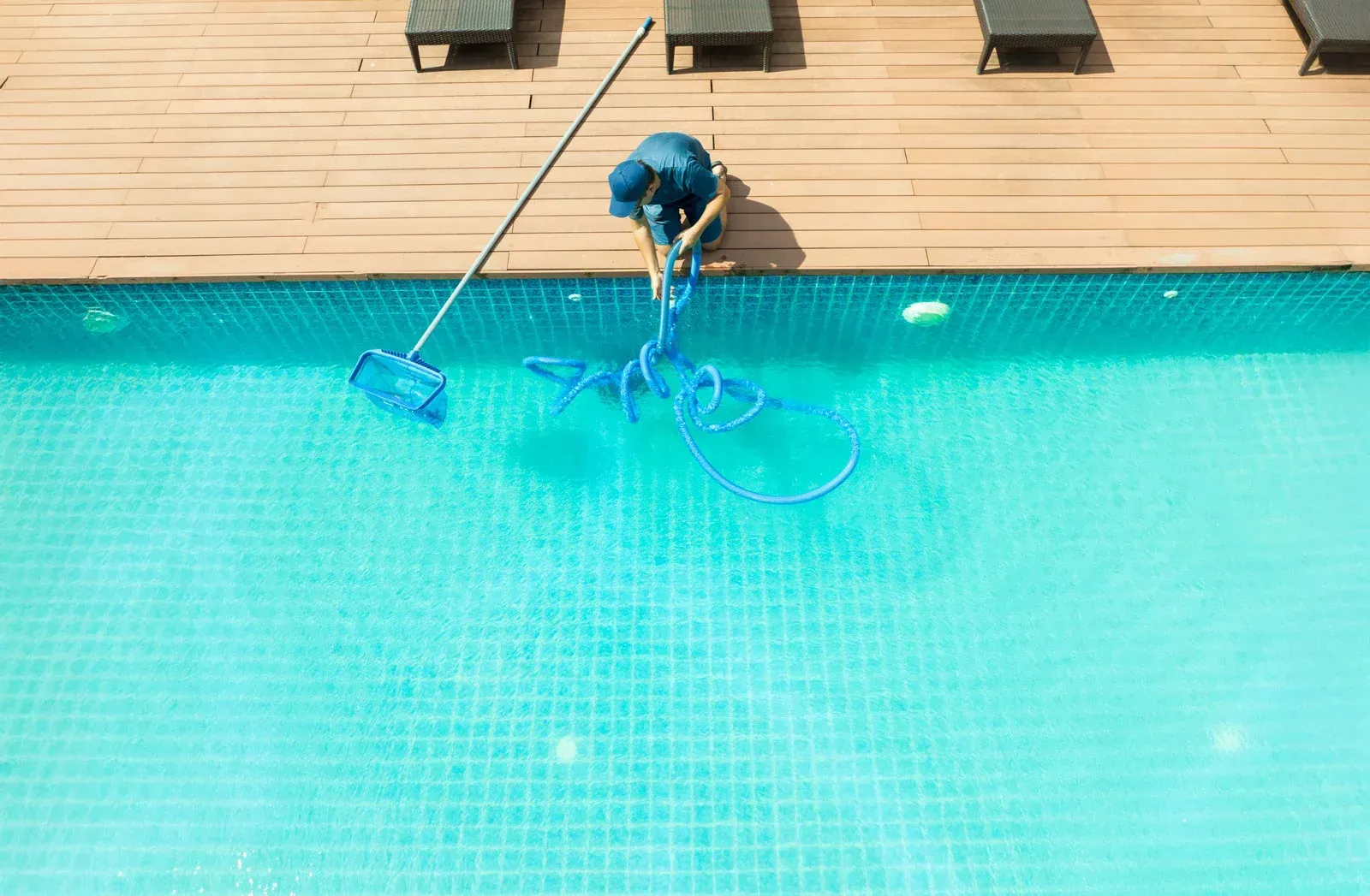 A person in blue cleaning a turquoise swimming pool with a long-handled net and vacuum hose from a wooden deck.