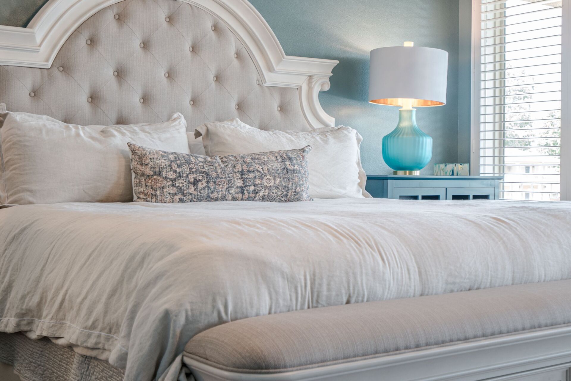 A bedroom with a bed , bench , lamp and window.