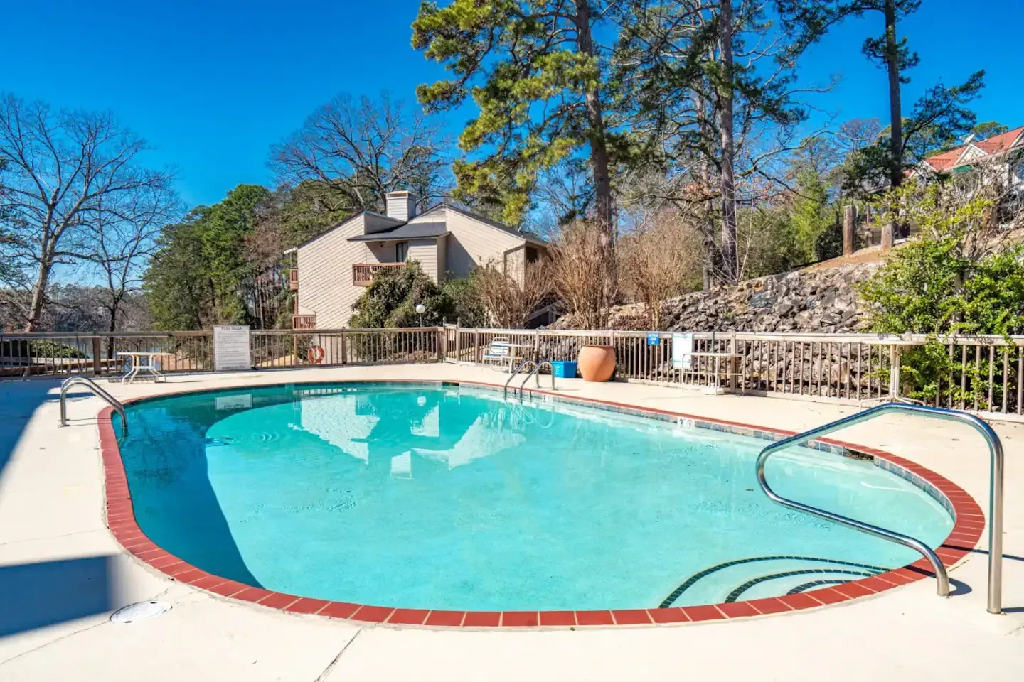 A large swimming pool is surrounded by a fence and trees in front of a house.