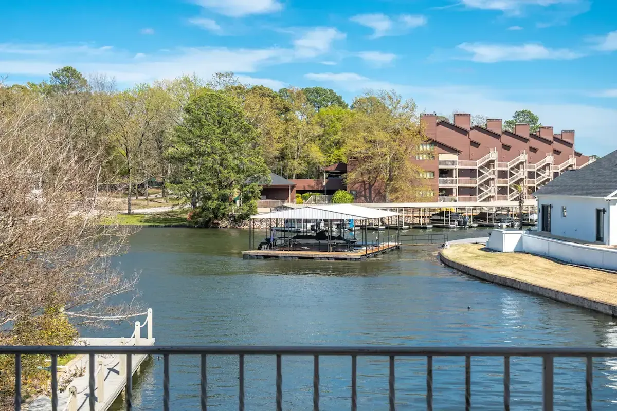 A view of a lake from a balcony with a dock and a house in the background.