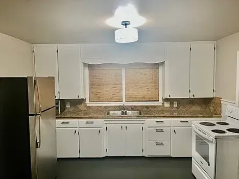 Kitchen with white cabinets, appliances, and a window with blinds; dark floor.
