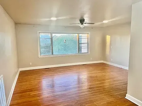 Empty living room with wood floors, large window, and ceiling fan.