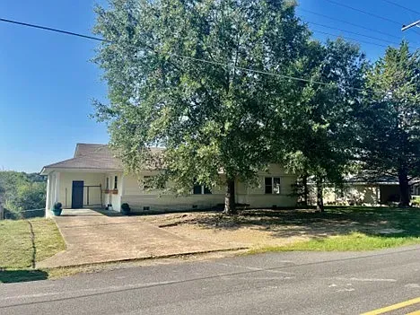 Beige house with a concrete driveway and large shade trees. Sunny day.
