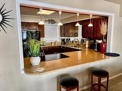 A kitchen with a bar and stools and a window between the kitchen and the living room.
