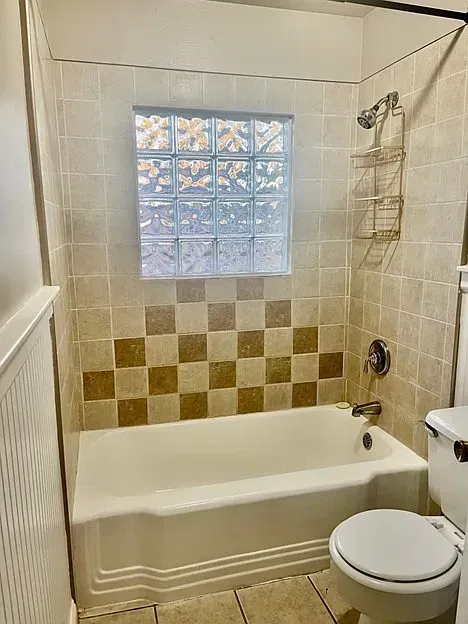 Bathroom with tub, beige tiles, brown checkerboard accent, glass block window, and toilet.