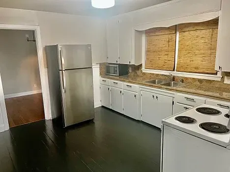 Kitchen with stainless steel refrigerator, white cabinets, and dark floor. Window has woven blinds.