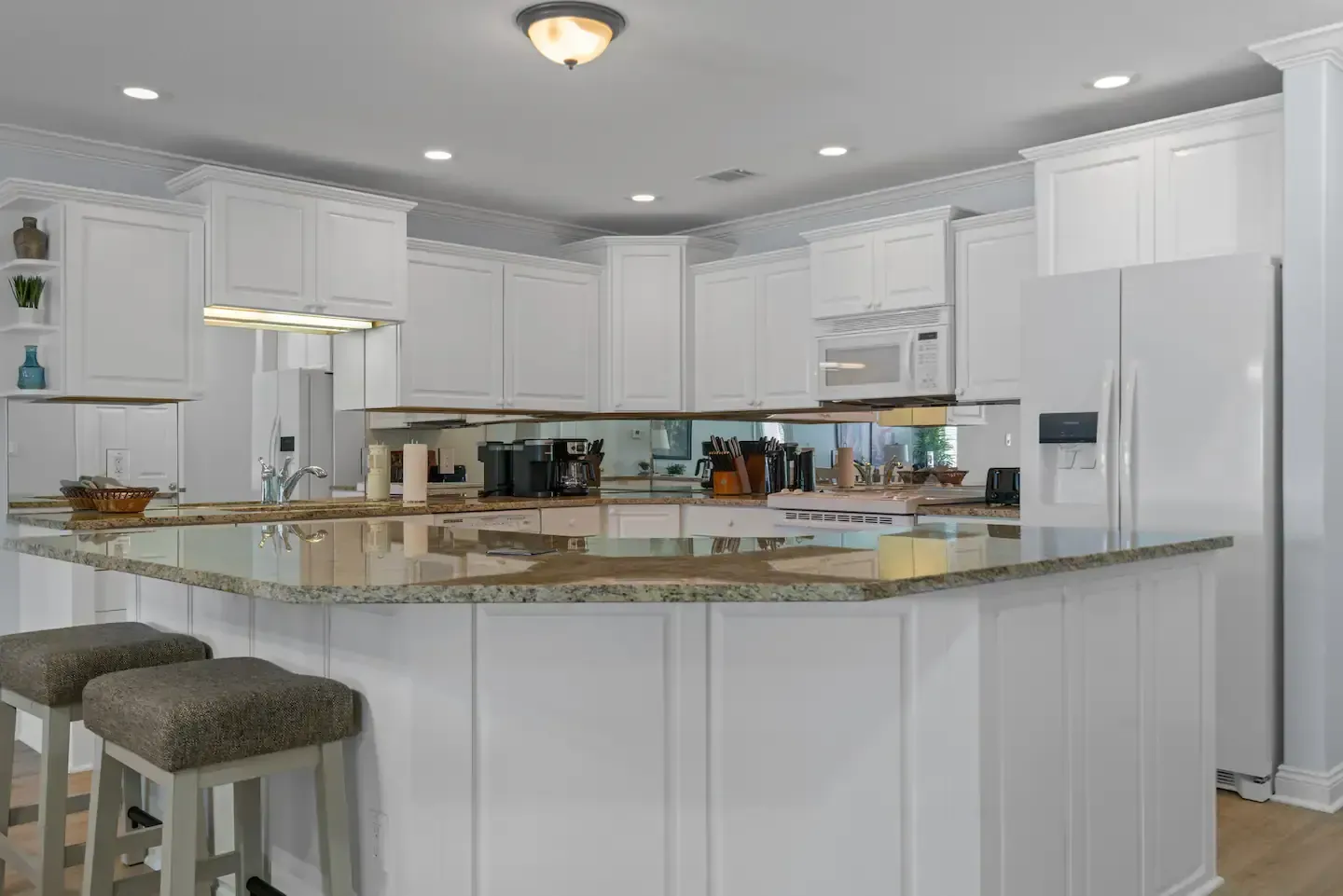 A kitchen with white cabinets , granite counter tops , stools and a refrigerator.