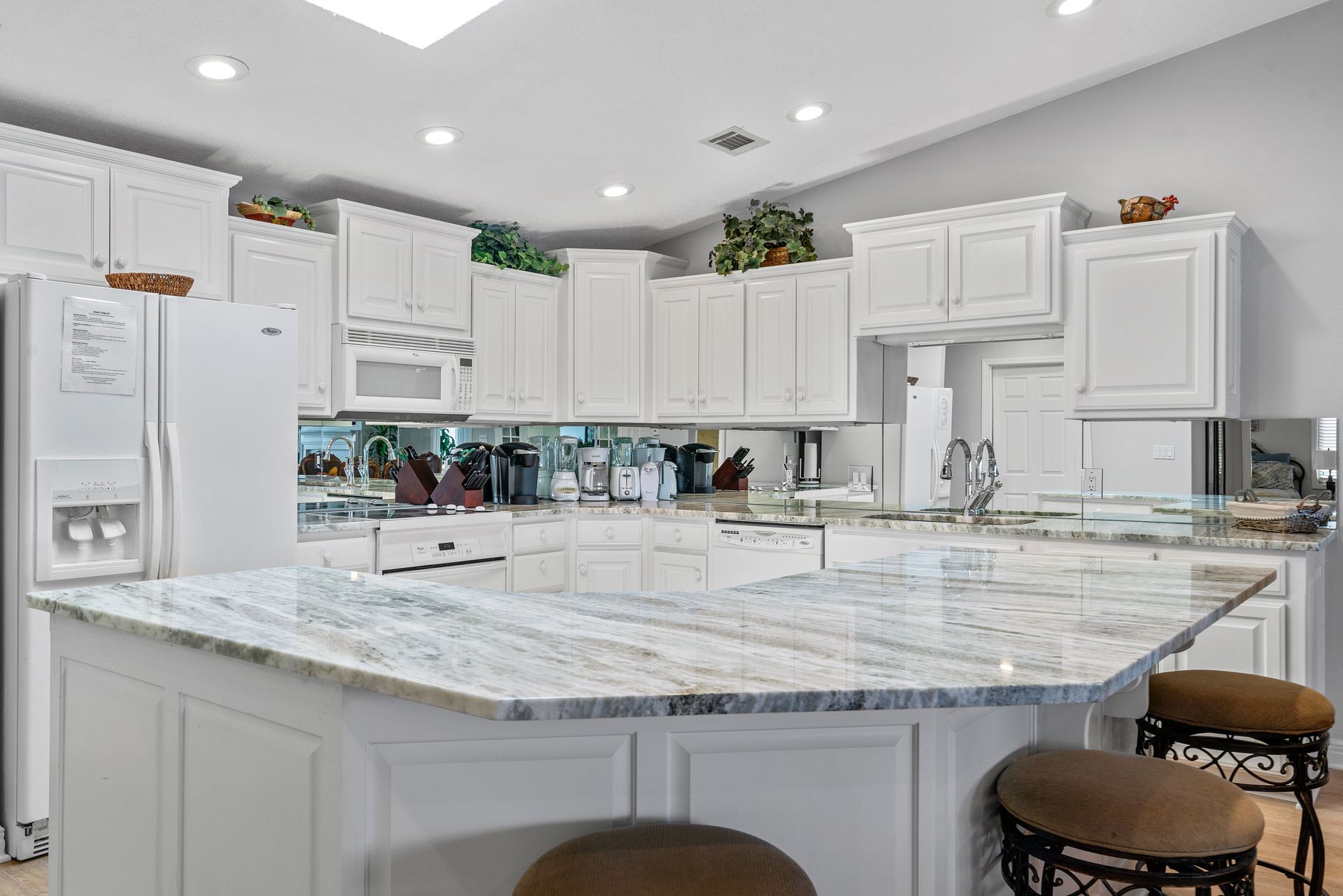 A kitchen with white cabinets , granite counter tops , stools and a refrigerator.
