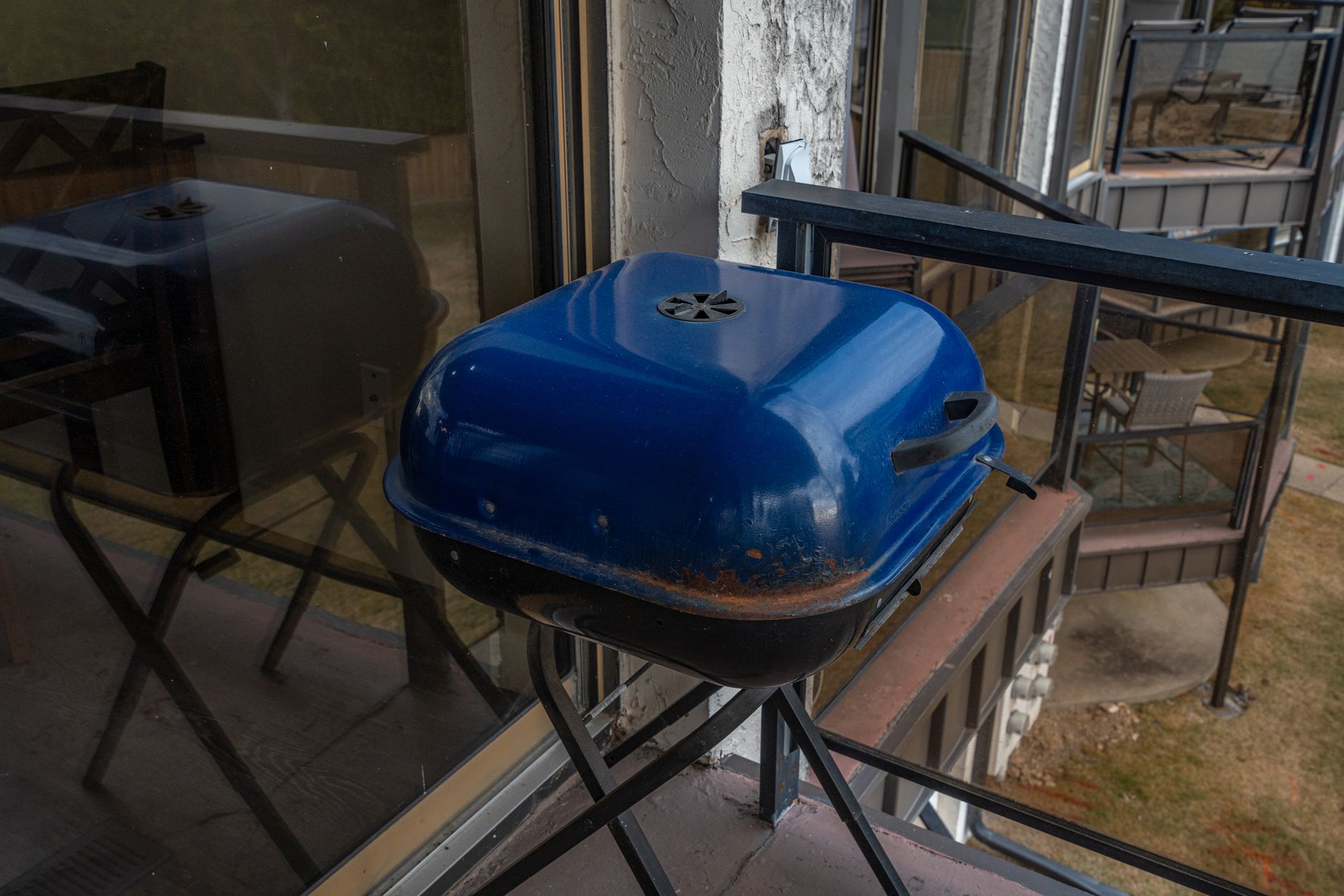 A blue grill is sitting on a balcony next to a window.
