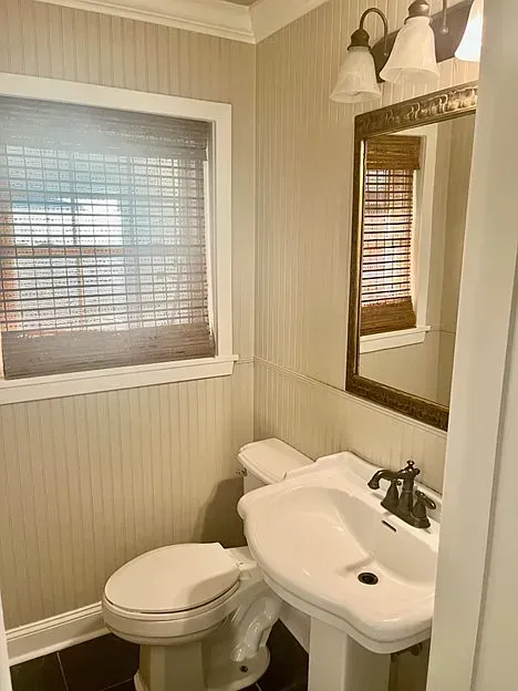 A small bathroom with beadboard walls, a toilet, pedestal sink, and window with a bamboo shade.