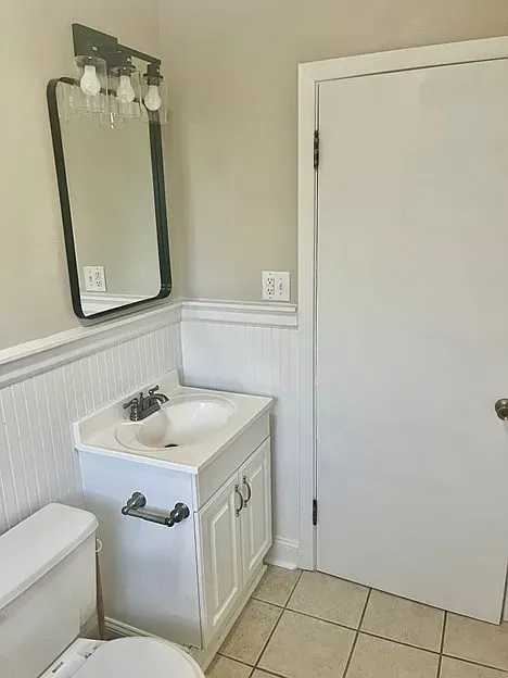Small bathroom with a white sink, toilet, and door; neutral walls, and a black-framed mirror.