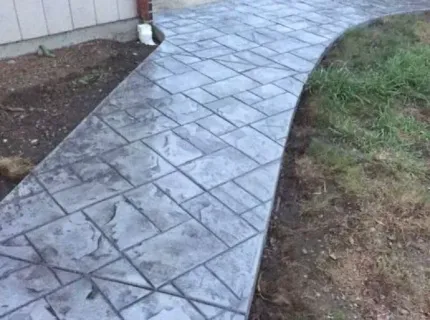 A concrete walkway leading to a house next to a grassy area.