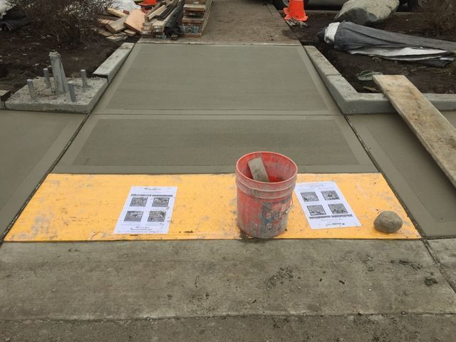 A bucket of concrete is sitting on a yellow mat on a sidewalk.