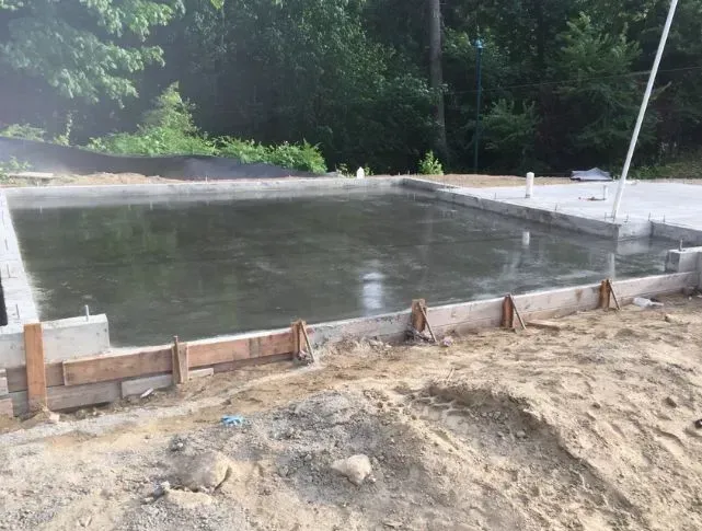 Newly poured concrete foundation within wooden forms, outdoors, surrounded by dirt and trees.
