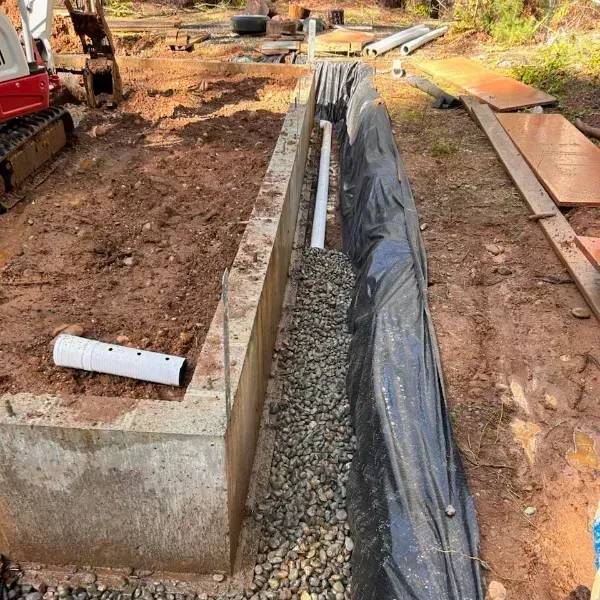 Trenches under construction with concrete walls, gravel, and black plastic sheeting beside excavated soil.
