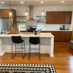 Modern kitchen with island, bar stools, and herringbone tile backsplash. Wood cabinets and floors.