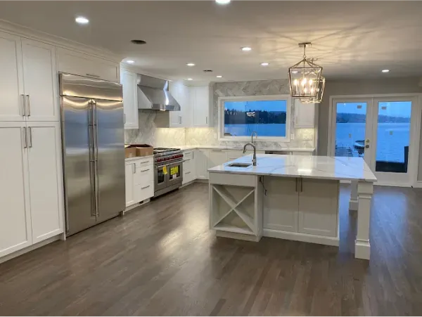 Spacious white kitchen with stainless steel appliances, marble countertops, and island, overlooking water.