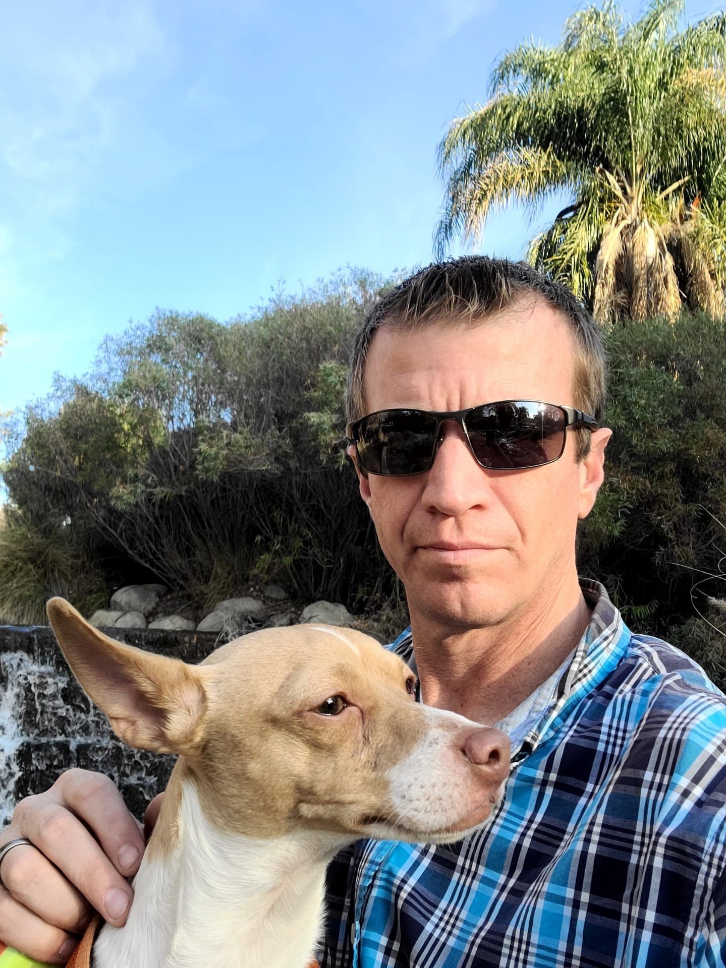 Man wearing sunglasses and a plaid shirt, smiling with a tan dog outdoors. Water and greenery visible.