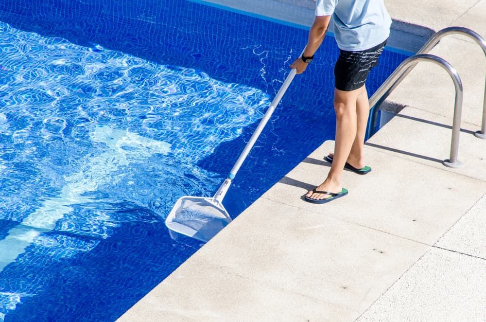 A Person in Flip-flops Uses a Pool Skimmer to Clean Debris From a Bright Blue Swimming Pool
