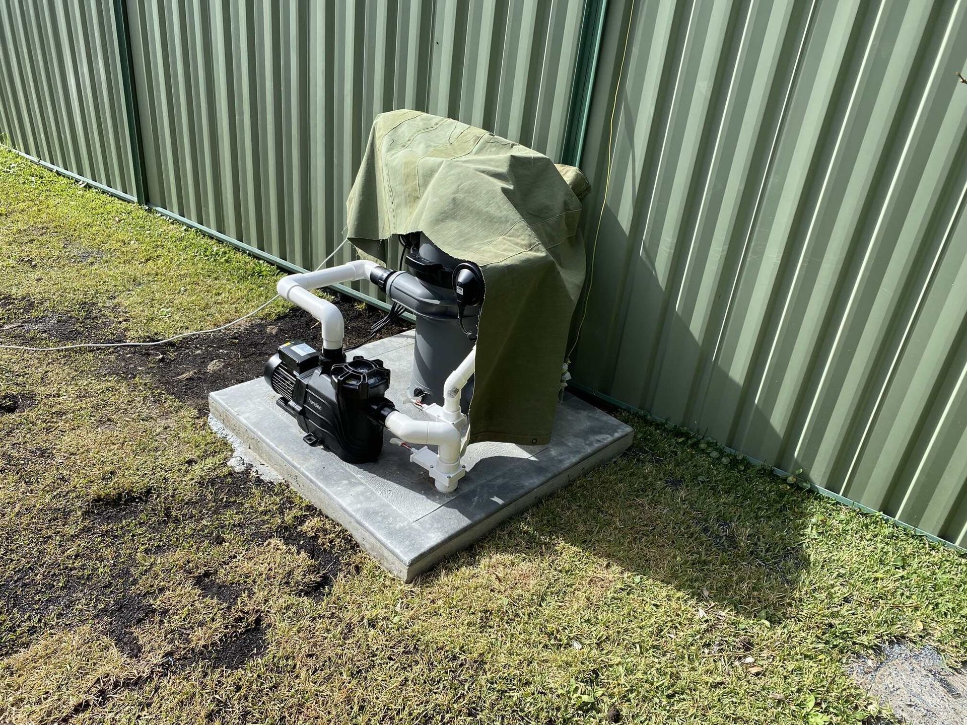 Pool filter system on a concrete pad, covered with a green tarp, beside a metal fence.