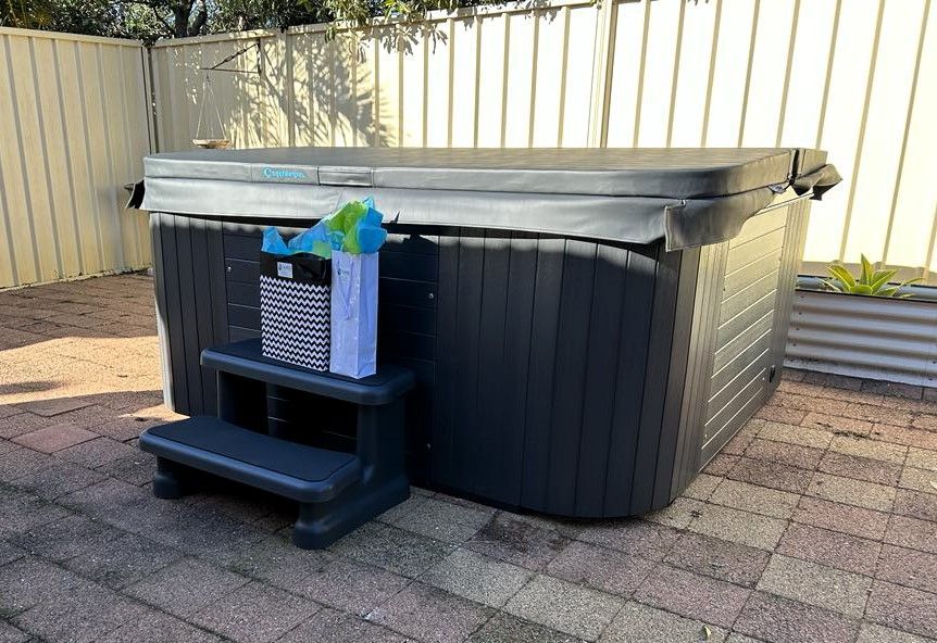 A Black Hot Tub is Sitting on a Brick Patio Next to a Fence — Taree Pool Supplies in Taree, NSW
