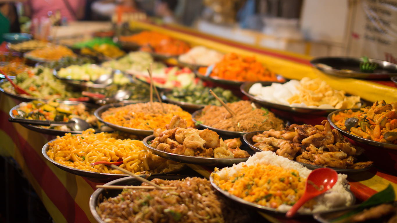 Platters of various prepared food items at an outdoor market, with yellow and orange accents.