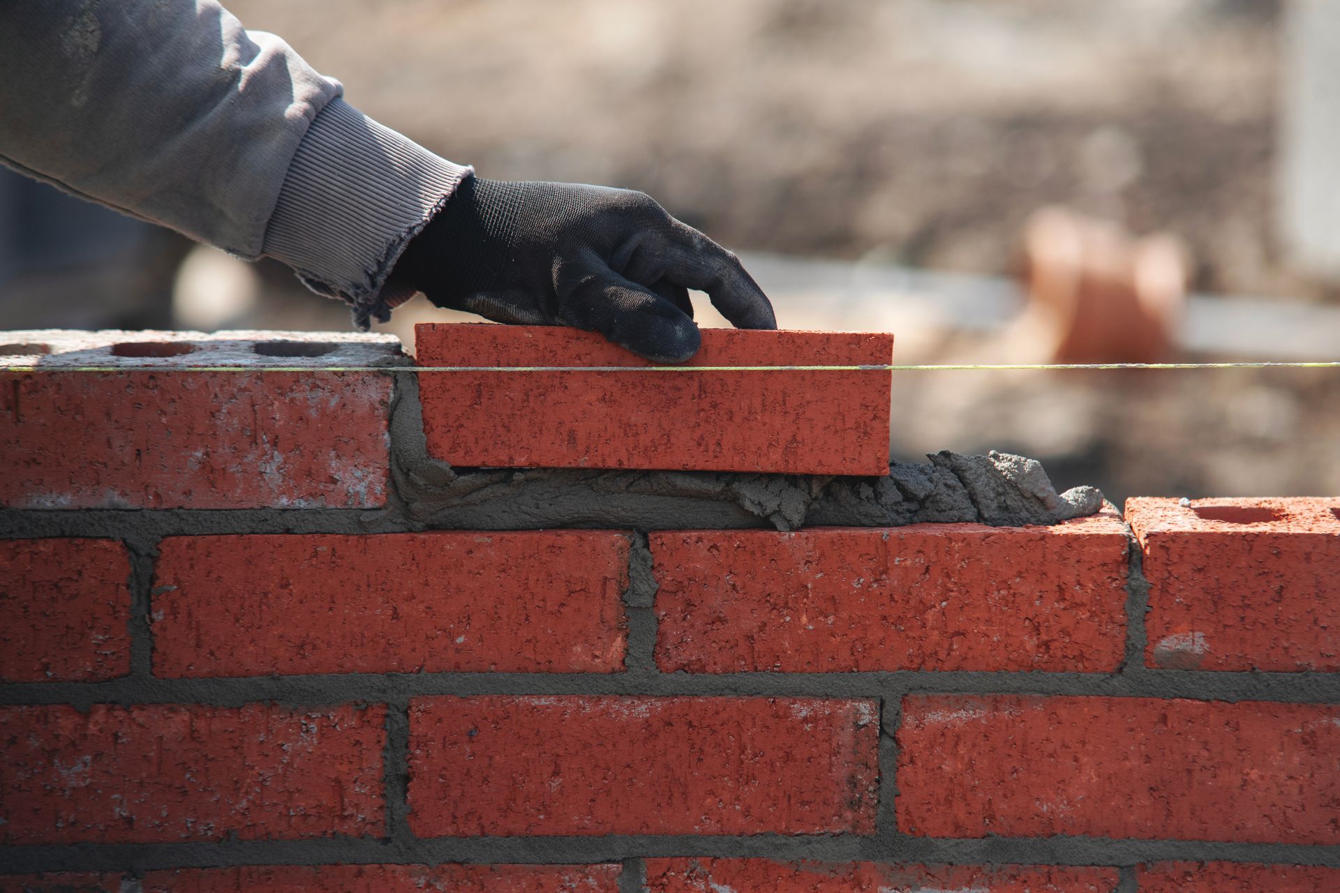 A gloved hand places a red brick onto a fresh bed of mortar atop a construction wall.