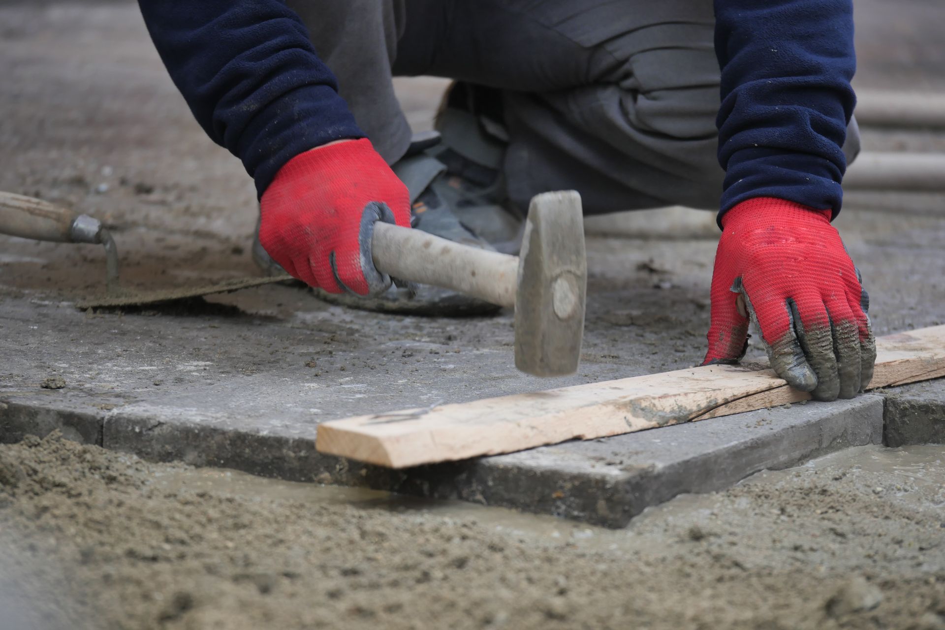 A worker wearing red gloves uses a hammer to tap a wooden board on top of newly laid stone pavers.