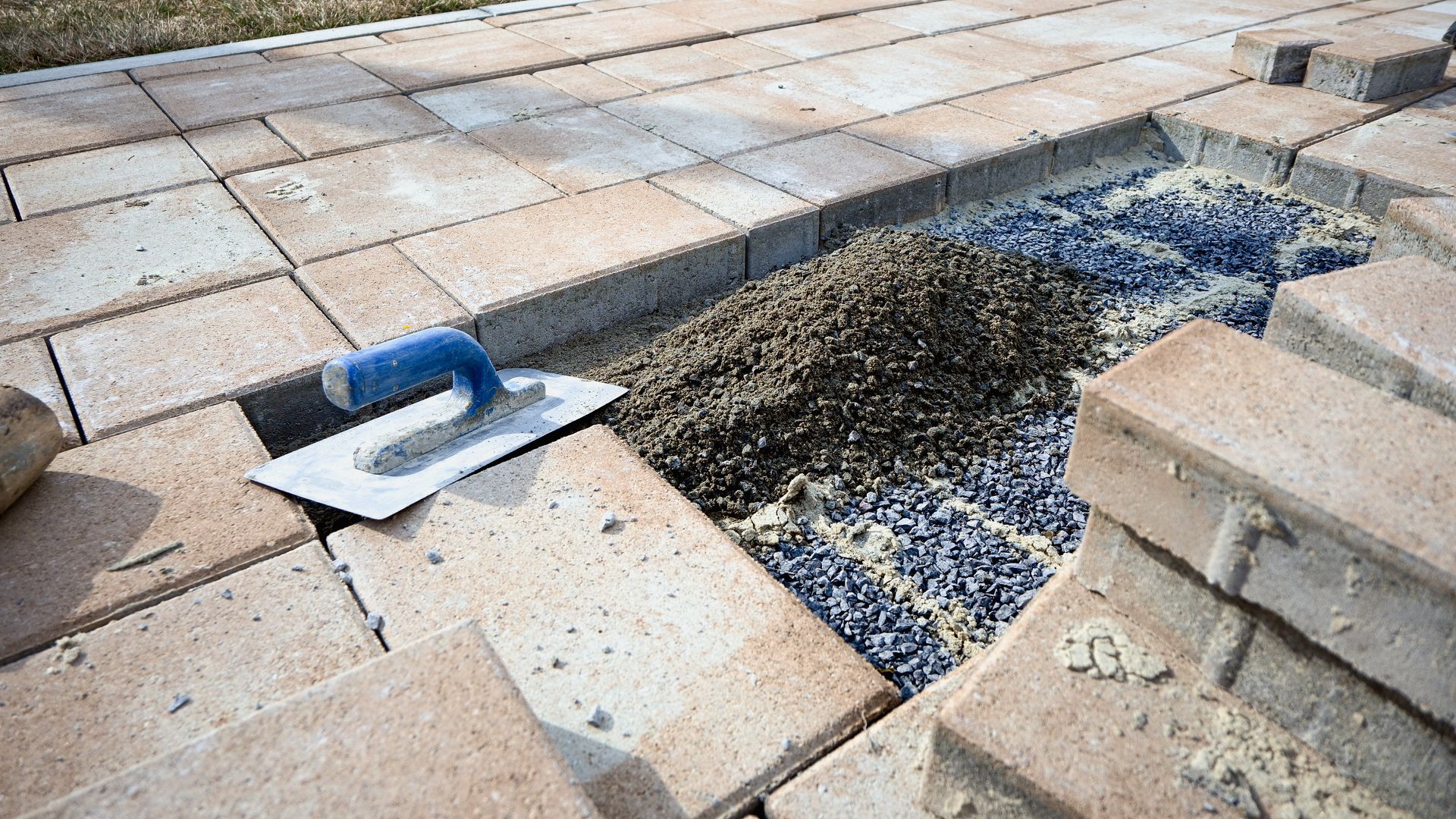 A metal trowel sits on tan paving stones next to a small section of unfinished, gravel-filled path under construction.
