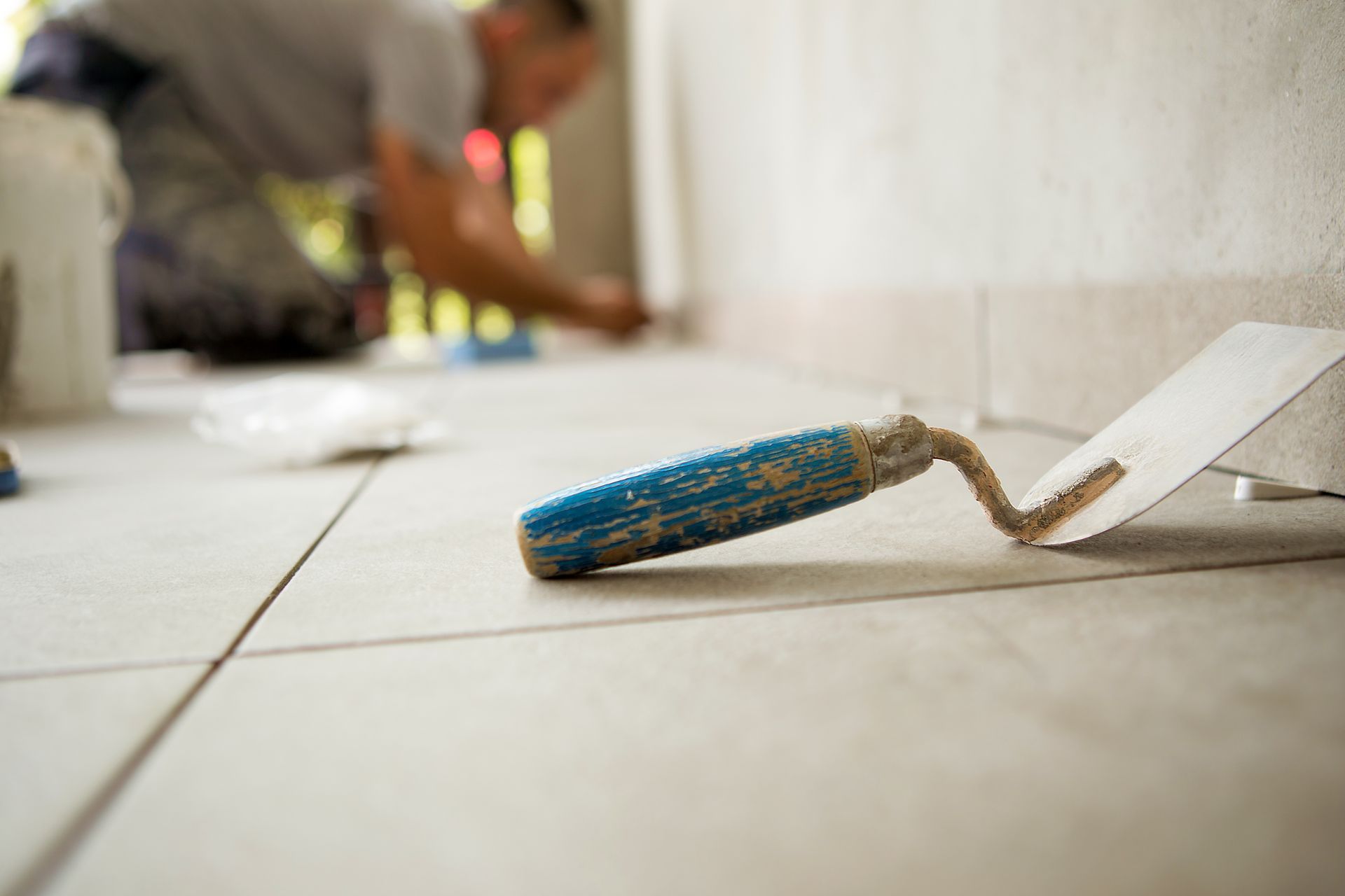 A close-up of a blue-handled trowel resting on newly laid light-colored tiles, with a person working in the background.