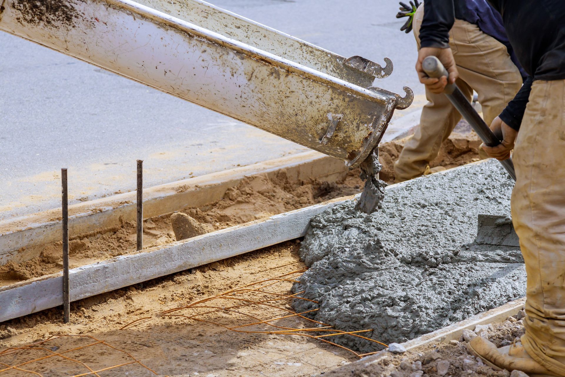 A worker uses a shovel to guide wet concrete pouring from a truck chute into a prepared form along a road.