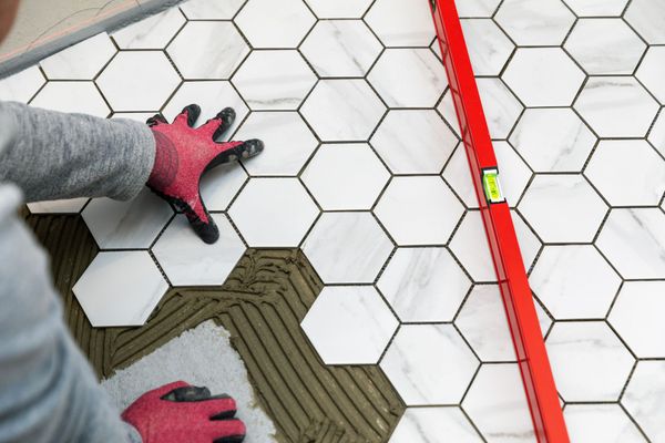 A gloved hand uses a trowel to apply mortar between white subway tiles on a wall, with tiling tools on a surface nearby.