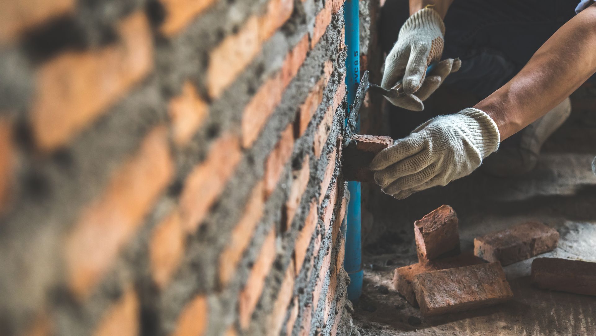 A construction worker in gloves lays a red brick into a mortar-filled wall next to a blue pipe.