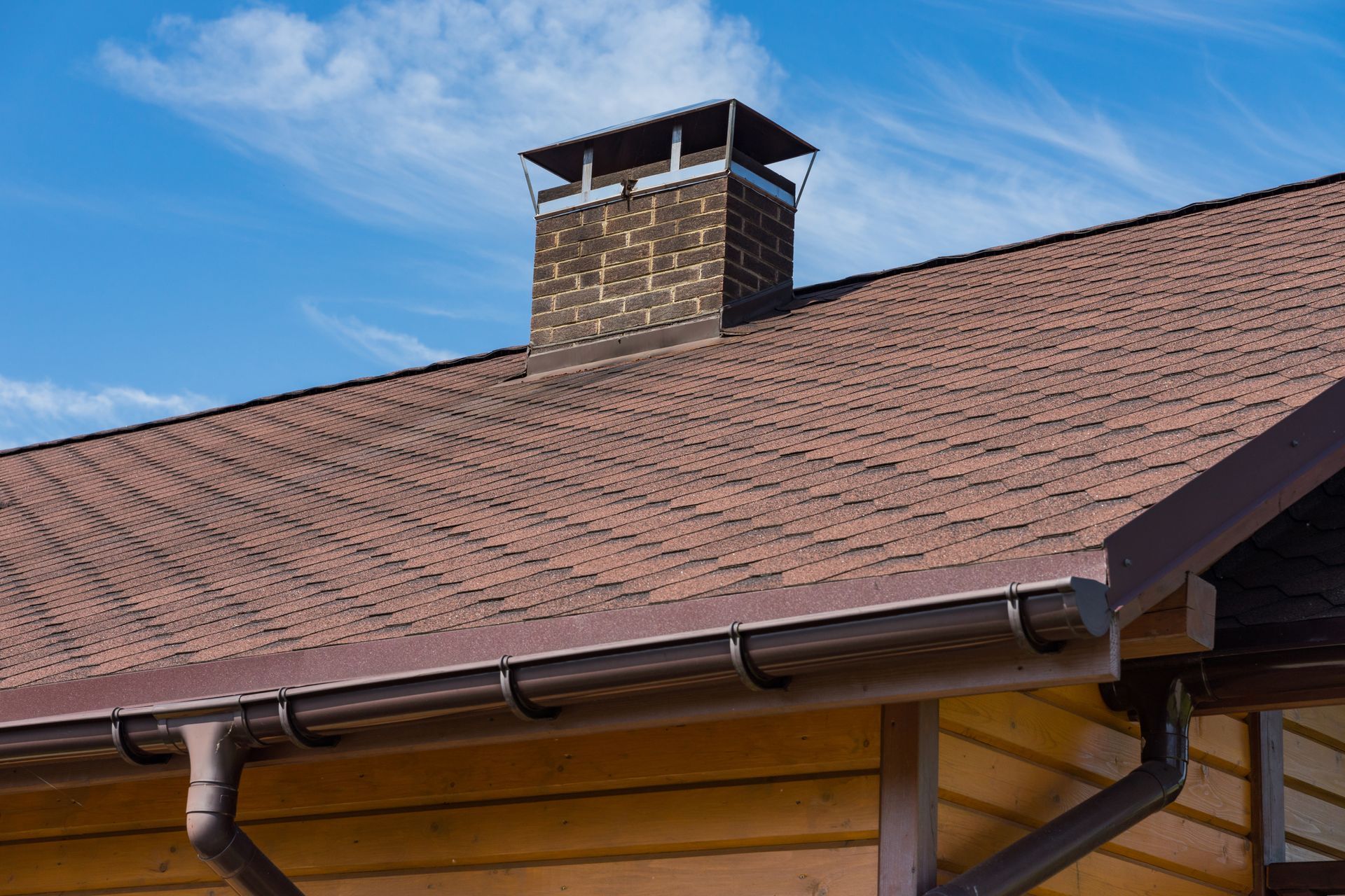 A brown shingled roof with a brick chimney and a gutter system against a blue sky.