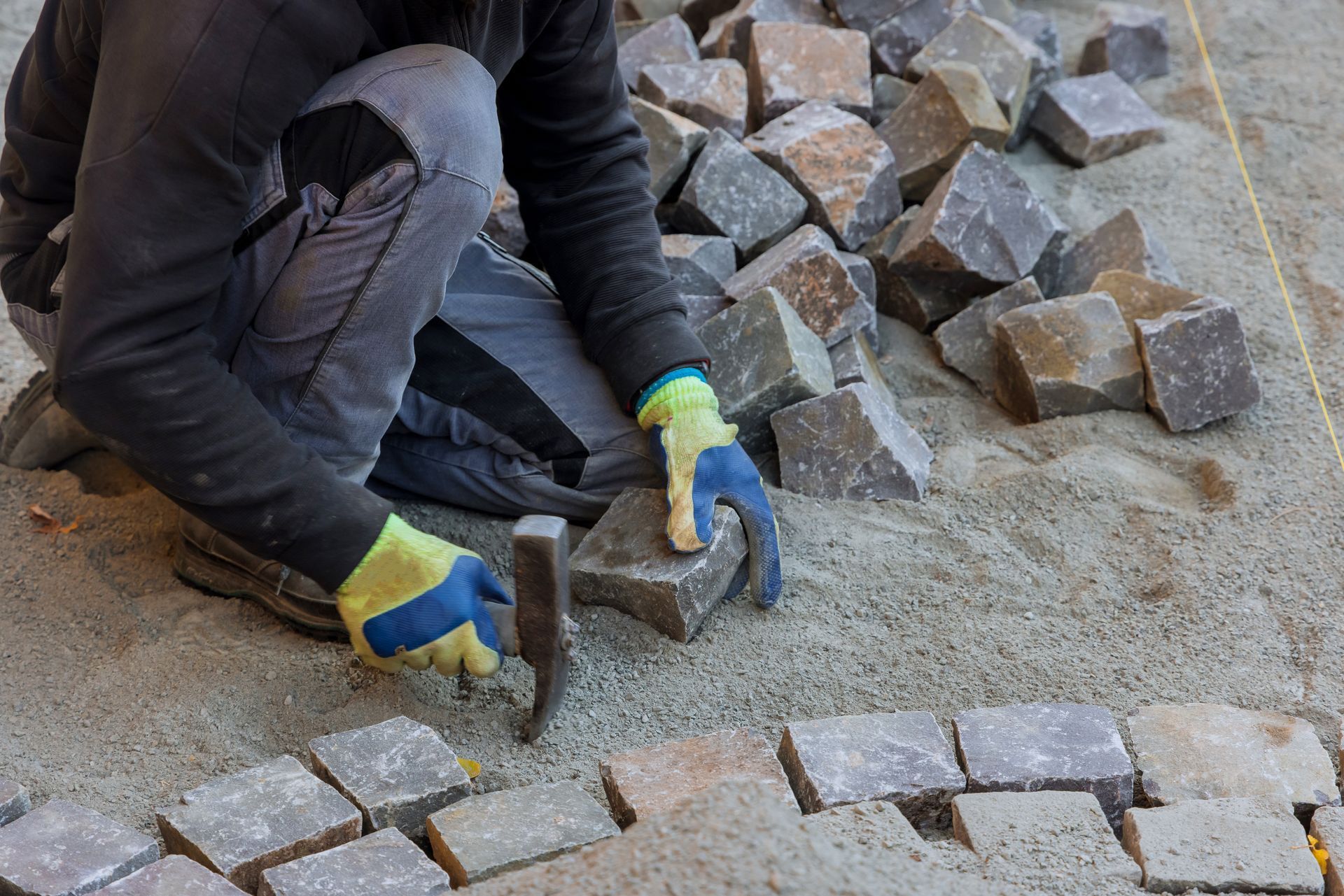 A worker wearing gloves kneels on sand, using a hammer to set stone pavers into a patterned walkway.