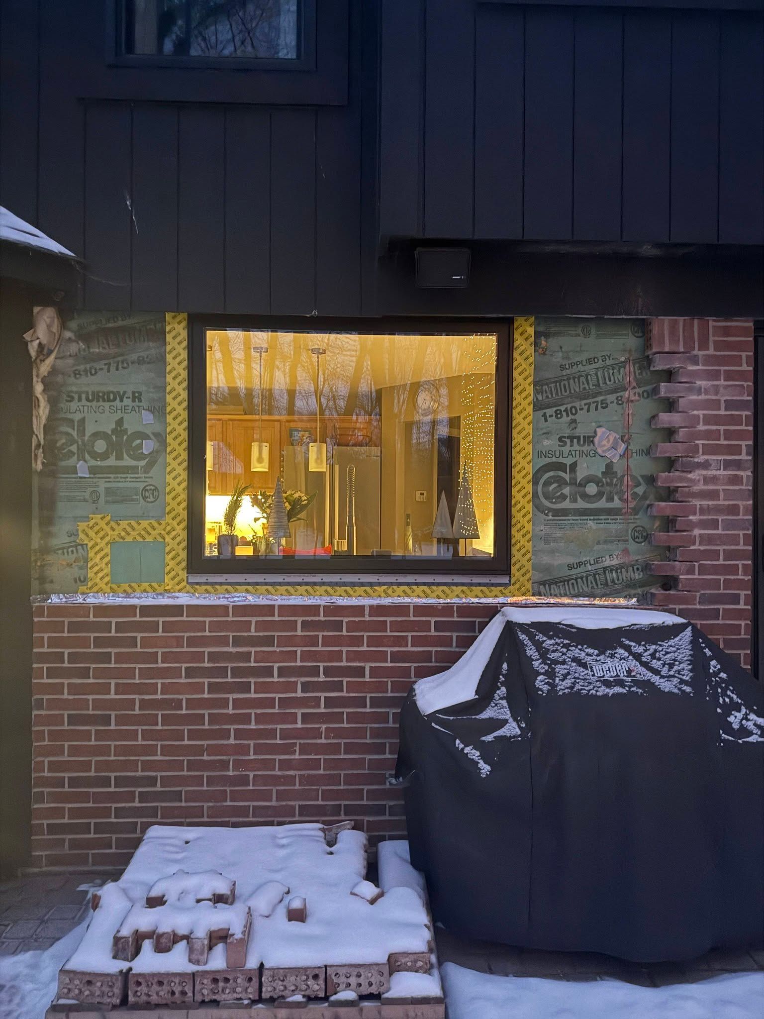 A house exterior featuring a window surrounded by green foam insulation panels, with a brick base and a snow-covered yard.
