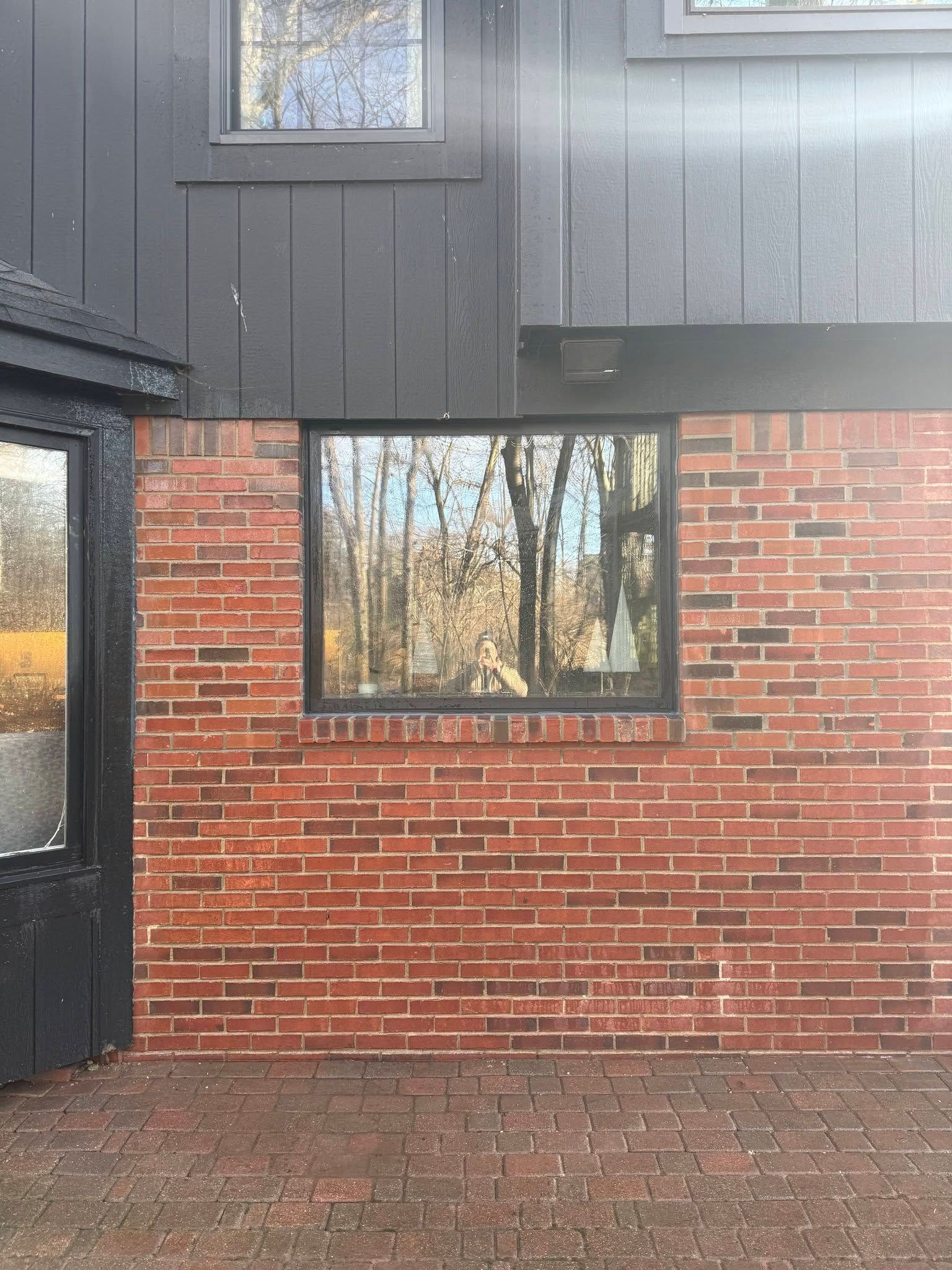 A brick exterior wall with a dark window reflecting trees and a black-paneled upper wall under a bright sky.