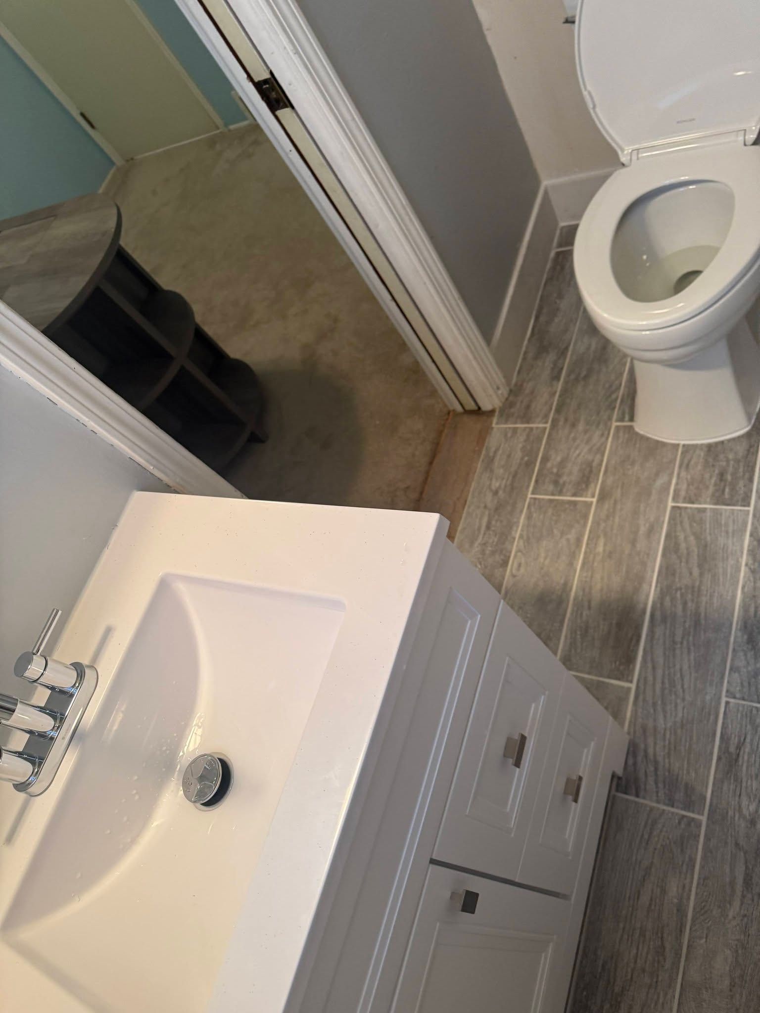 A high-angle shot of a bathroom featuring a white vanity with a sink, grey wood-look tile flooring, and a white toilet.