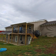 A multi-level home with a tan exterior, featuring an upper deck with wood posts and a lower covered patio on a grassy hill.