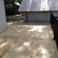 A newly built wooden deck with black metal railings, situated next to a house wall and a white building with a tin roof.
