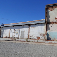 A weathered, white industrial building with exposed brick trim and a gravel foreground under a bright blue sky.