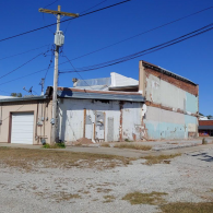 A dilapidated, weathered building with a garage door and exposed brick under a clear blue sky, next to a utility pole.