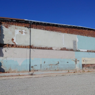 An old brick and stucco wall in disrepair with patches of peeling blue and white paint, set against a clear blue sky.