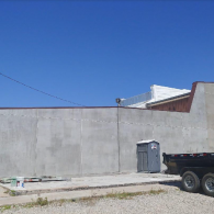 A concrete wall stands under a clear blue sky, next to a portable toilet and a construction trailer on a gravel lot.